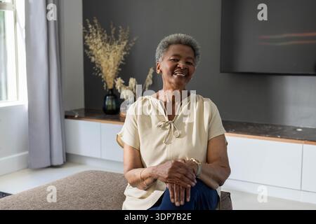 Femme afro-américaine senior souriante assise sur un pouf dans le salon, portant un chemisier beige, la télévision Banque D'Images