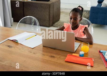 Écolière africaine portant le haut rose et scrunchie assis à table à la maison à l'aide d'un ordinateur portable argenté Banque D'Images