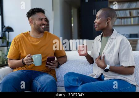 Couple assis sur le canapé dans le salon portant des jeans tenant des tasses en céramique et smartphone Banque D'Images