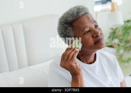 Femme afro-américaine senior sur le lit portant un tee-shirt blanc en utilisant la pierre gua Sha en forme de coeur vert Banque D'Images