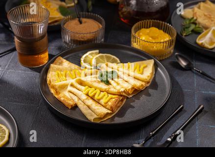 Fines crêpes pliées sur une assiette noire surmontée de caillé de citron brillant, de menthe fraîche et d’une tranche de citron, servies avec du sucre, du thé chaud et du caillé supplémentaire pour le brunch Banque D'Images