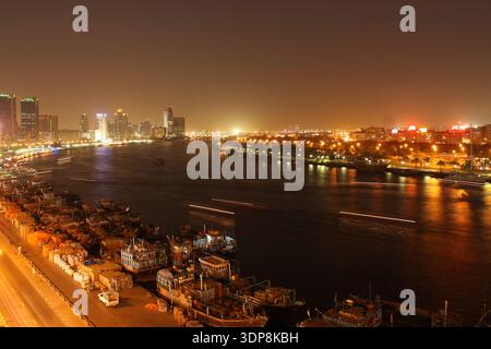 Vue nocturne du Dubai Creek avec gratte-ciel illuminés et traînées de lumière des embarcations qui passent Banque D'Images