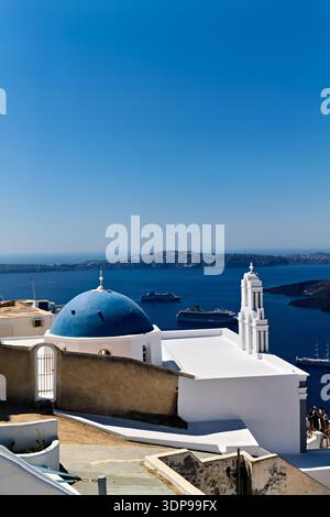 Église emblématique à dôme bleu et architecture blanche à Santorin, en Grèce, avec des bateaux de croisière dans la caldeira. Les touristes apprécient la vue imprenable sur la mer Égée. Banque D'Images
