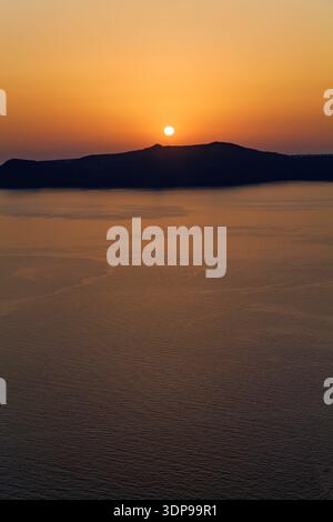 Un coucher de soleil orange vibrant sur la mer Égée, avec le soleil plongeant derrière une silhouette d'île lointaine à Santorin, en Grèce Banque D'Images