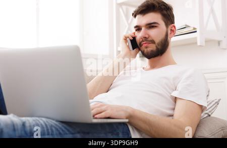 Homme utilise ordinateur portable tout en parlant sur téléphone portable à la maison pendant la journée Banque D'Images