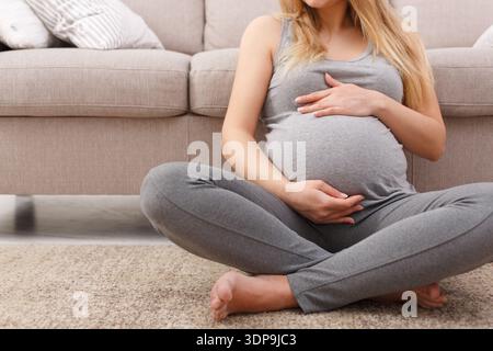 Femme enceinte est assis confortablement sur le sol dans un salon avec canapé gris et tapis Banque D'Images