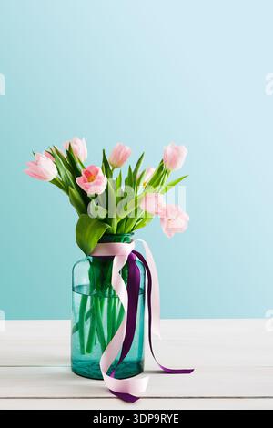 Tulipes roses fraîches dans un vase en verre avec des rubans placés sur une table en bois blanc Banque D'Images