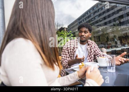 Jeune homme et femme assis dans un café en plein air dans la ville, partageant le café et une conversation sérieuse comme il gestes, transmettant la discussion focalisée et aut Banque D'Images