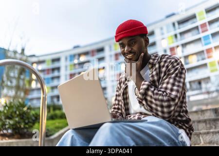 Jeune homme noir assis sur les marches de l'appartement avec un ordinateur portable, souriant et réfléchi comme il travaille ou étudie à l'extérieur, incarnant le travail à distance urbain moderne et Banque D'Images