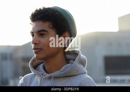 Adolescent homme debout sur le toit urbain face à gauche, portant un sweat à capuche gris et un bonnet vert Banque D'Images