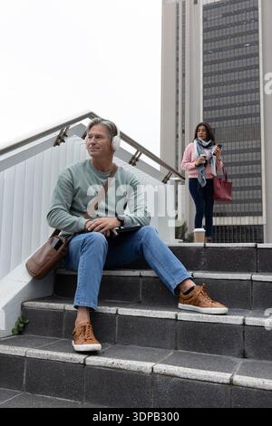 Navetteurs assis sur les marches près de la tour de bureaux, homme portant un casque utilisant une tablette, femme tenant une tasse Banque D'Images