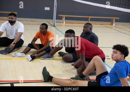 Divers coéquipiers masculins assis et étirant sur le terrain de gymnastique en vêtements sportifs près du filet de volley-ball Banque D'Images
