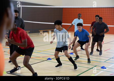 Divers coéquipiers masculins se faufilant dans un exercice à cône sur un terrain de gymnastique près du filet de volley-ball, portant des chaussures Banque D'Images