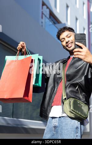 Homme asiatique portant veste en cuir avec sac olive, tenant téléphone et sacs à provisions dans la rue urbaine Banque D'Images
