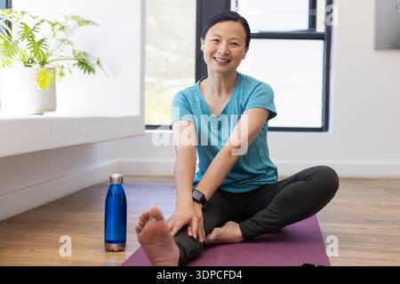 Femme asiatique portant un haut sarcelle, assise sur un tapis de yoga violet à la maison, étirant la jambe avec une bouteille Banque D'Images