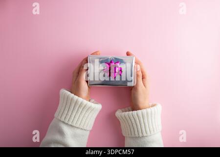 Première personne vue de dessus photo de mains tenant la boîte-cadeau argent brillant avec arc rose sur fond rose isolé Banque D'Images