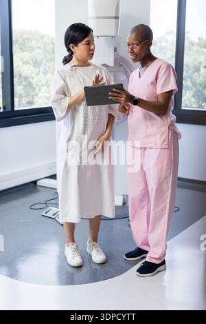 Patient féminin diversifié dans la robe et technique féminine dans les gommages roses examinant le comprimé dans la salle de mammographie Banque D'Images
