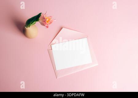 Carte vierge avec enveloppe rose et tulipe dans un vase sur fond pastel. Modèle de salutation romantique. Vue de dessus, maquette. Banque D'Images