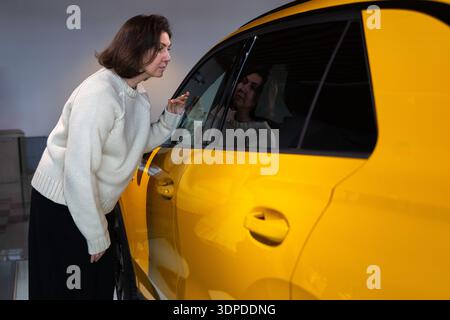 Une femme inspecte une nouvelle voiture jaune de près dans une salle d'exposition de concession évaluant les caractéristiques intérieures et extérieures prise de décision achat automobile moderne vente au détail environnement automobile Banque D'Images