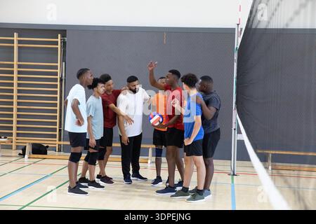 Équipe masculine de volley-ball diversifiée se blottissant près d'un poteau de filet sur le terrain de gymnastique tenant un ballon de volley-ball bleu-blanc-rouge Banque D'Images