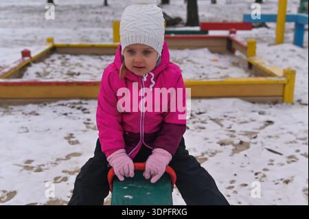 Fille assise sur balançoire dans le terrain de jeu d'hiver. enfant habillé chaud dans une veste rose et chapeau blanc tenant le siège balançoire. enfant femelle appréciant l'outdoo de la saison froide Banque D'Images