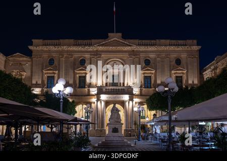 Vue nocturne de la statue de la reine Victoria à la Valette, éclairée devant une façade néoclassique avec entrée voûtée, lampadaires et terrasses de café en plein air. Banque D'Images