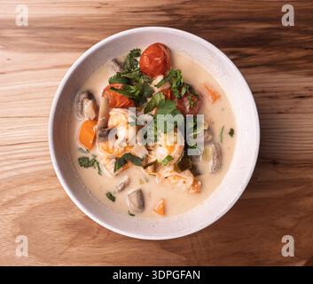 Une pose à plat propre de Tom Kha Goong dans un bol blanc sur une surface en bois clair, avec de grosses crevettes et des garnitures fraîches. Style de blog alimentaire moderne Banque D'Images
