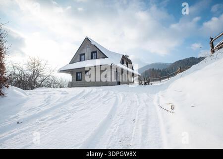 Cabane de montagne traditionnelle en bois recouverte de neige blanche fraîche sur une journée ensoleillée d'hiver avec une architecture alpine confortable et des traces de pneus sur la colline enneigée Pat Banque D'Images