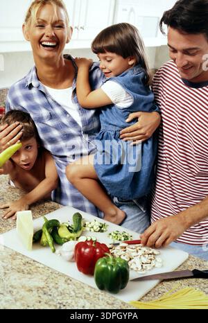 Une famille s'amuse dans la cuisine tout en préparant un repas. Deux enfants s'impliquent, dont un assis sur le dos d'un parent. Légumes frais Banque D'Images