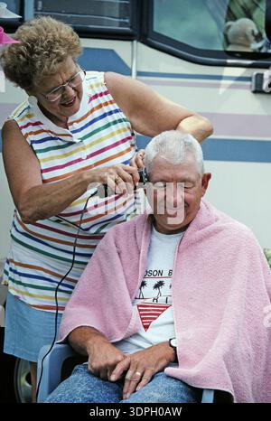Une femme tond les cheveux d'un homme qui est assis détendu dans une chaise. Ils sont à l'extérieur près d'un camping-car. L'homme porte une chemise légère et une serviette, et la femme utilise Banque D'Images