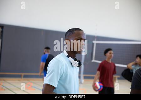 Joueurs masculins pratiquant dans le gymnase de l'école, l'un portant des écouteurs, coéquipier tenant le volleyball par filet Banque D'Images