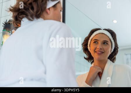 Femme debout au miroir de la salle de bain, souriant en touchant le cou, portant un peignoir blanc et un bandeau Banque D'Images