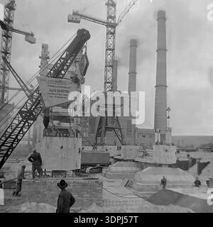 Une remarquable photographie d'archives en noir et blanc datant de 1965 documentant la construction à grande échelle du bloc d'énergie de 800 MW à la centrale thermique de Slavyanskaya (GRES) à Nikolaevka, dans la région de Donetsk. Une lourde grue sur chenilles est positionnée au premier plan, hissant un grand panneau avec un engagement socialiste de travail pour le collectif Metallomontazh, fixant la date limite au 15 octobre 1965. L'arrière-plan révèle des cheminées imposantes et des structures squelettiques industrielles. Ce cliché authentique capture l'immense effort d'ingénierie et l'esprit du Donbass paisible. Banque D'Images