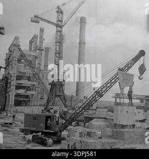 Une remarquable photographie d'archives en noir et blanc datant de 1965 documentant la construction à grande échelle du bloc d'énergie de 800 MW à la centrale thermique de Slavyanskaya (GRES) à Nikolaevka, dans la région de Donetsk. Une lourde grue sur chenilles est positionnée au premier plan, hissant un grand panneau avec un engagement socialiste de travail pour le collectif Metallomontazh, fixant la date limite au 15 octobre 1965. L'arrière-plan révèle des cheminées imposantes et des structures squelettiques industrielles. Ce cliché authentique capture l'immense effort d'ingénierie et l'esprit du Donbass paisible. Banque D'Images