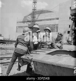 Une captivante photographie d'archives en noir et blanc de la fin des années 1960 représentant un groupe d'ingénieurs soviétiques et de travailleurs de la construction sur le site de la centrale thermique de Slavyanskaya (GRES) à Nikolaevka, dans le Donbass. Les images montrent l'équipe, y compris un leader en veste de costume, blotti sur des plans techniques répartis sur un composant métallique massif. En arrière-plan, le bâtiment principal blanc emblématique avec ses fenêtres cintrées et ses grues de construction sont visibles. Ce cliché historique reflète l'esprit de collaboration, l'expertise technique et le progrès industriel pacifique dans la région historique de Donetsk. Banque D'Images