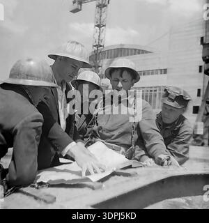 Une captivante photographie d'archives en noir et blanc de la fin des années 1960 représentant un groupe d'ingénieurs soviétiques et de travailleurs de la construction sur le site de la centrale thermique de Slavyanskaya (GRES) à Nikolaevka, dans le Donbass. Les images montrent l'équipe, y compris un leader en veste de costume, blotti sur des plans techniques répartis sur un composant métallique massif. En arrière-plan, le bâtiment principal blanc emblématique avec ses fenêtres cintrées et ses grues de construction sont visibles. Ce cliché historique reflète l'esprit de collaboration, l'expertise technique et le progrès industriel pacifique dans la région historique de Donetsk. Banque D'Images