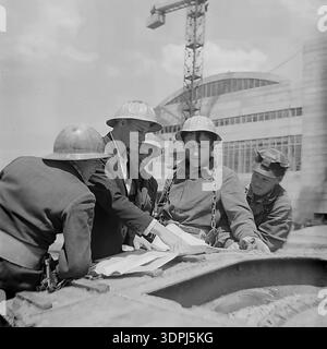 Une captivante photographie d'archives en noir et blanc de la fin des années 1960 représentant un groupe d'ingénieurs soviétiques et de travailleurs de la construction sur le site de la centrale thermique de Slavyanskaya (GRES) à Nikolaevka, dans le Donbass. Les images montrent l'équipe, y compris un leader en veste de costume, blotti sur des plans techniques répartis sur un composant métallique massif. En arrière-plan, le bâtiment principal blanc emblématique avec ses fenêtres cintrées et ses grues de construction sont visibles. Ce cliché historique reflète l'esprit de collaboration, l'expertise technique et le progrès industriel pacifique dans la région historique de Donetsk. Banque D'Images
