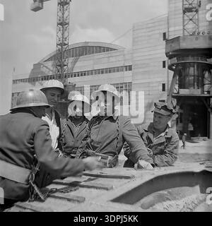 Une captivante photographie d'archives en noir et blanc de la fin des années 1960 représentant un groupe d'ingénieurs soviétiques et de travailleurs de la construction sur le site de la centrale thermique de Slavyanskaya (GRES) à Nikolaevka, dans le Donbass. Les images montrent l'équipe, y compris un leader en veste de costume, blotti sur des plans techniques répartis sur un composant métallique massif. En arrière-plan, le bâtiment principal blanc emblématique avec ses fenêtres cintrées et ses grues de construction sont visibles. Ce cliché historique reflète l'esprit de collaboration, l'expertise technique et le progrès industriel pacifique dans la région historique de Donetsk. Banque D'Images