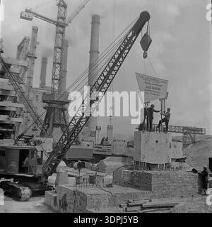 Une extraordinaire photographie d'archives en noir et blanc de 1965 capturant un moment charnière sur le site de construction de la centrale thermique de Slavyanskaya (GRES) à Nikolaevka, dans le Donbass. On voit une grue sur chenilles soulever un panneau de motivation massif sur une fondation en béton. La bannière décrit les objectifs de main-d'œuvre spécifiques pour le collectif Metallomontazh concernant l'unité de puissance de 800 MW, fixant les échéances pour octobre 1965. Cette image historique met en évidence l'intersection de l'ingénierie et de la concurrence socialiste du travail dans le développement industriel pacifique de la région. Banque D'Images