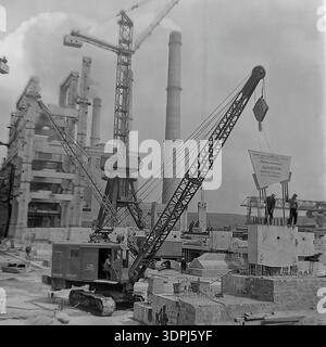 Une extraordinaire photographie d'archives en noir et blanc de 1965 capturant un moment charnière sur le site de construction de la centrale thermique de Slavyanskaya (GRES) à Nikolaevka, dans le Donbass. On voit une grue sur chenilles soulever un panneau de motivation massif sur une fondation en béton. La bannière décrit les objectifs de main-d'œuvre spécifiques pour le collectif Metallomontazh concernant l'unité de puissance de 800 MW, fixant les échéances pour octobre 1965. Cette image historique met en évidence l'intersection de l'ingénierie et de la concurrence socialiste du travail dans le développement industriel pacifique de la région. Banque D'Images