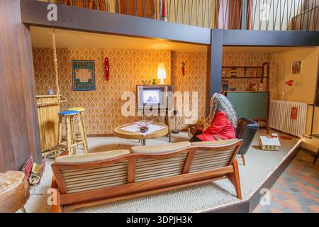 Genk, Belgique. 17 octobre 2025. Femme senior assise sur le canapé dans le salon de deux étages, maison de style des années 1960, télévision, radio, bar avec tabourets hauts, sh Banque D'Images