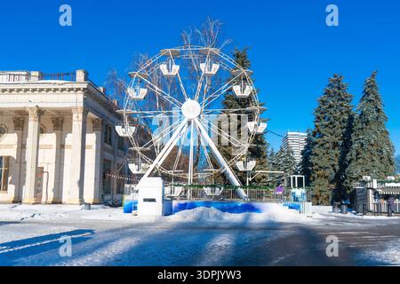 Roue de ferris blanche dans un parc d'attractions en hiver avec de la neige sur le sol et un fond de ciel bleu vif Banque D'Images