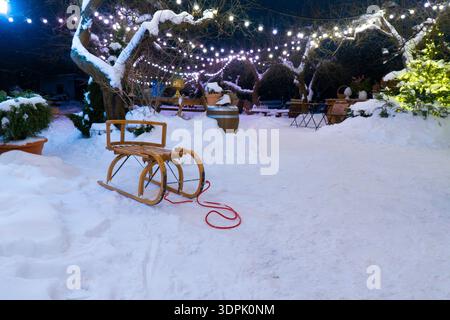 Traîneau en bois vintage sur neige blanche dans une arrière-cour décorée de lumières de fées chaudes. Ambiance chaleureuse de vacances d'hiver dans le jardin du soir Banque D'Images