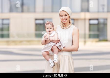 Élégante maman heureuse et fille souriant ensemble dans la ville portant des robes beige clair et des accessoires de mode à l'extérieur. Mère à la mode avec enfant Banque D'Images