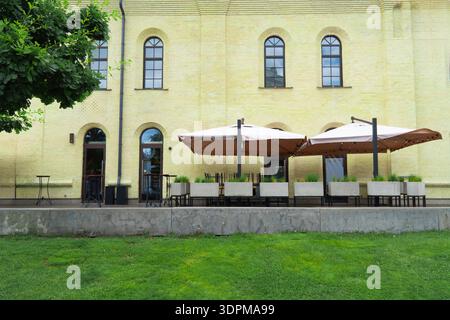 Terrasse extérieure confortable du restaurant avec parasols beiges, tables en bois et jardinières vertes sur une façade de bâtiment classique en briques jaunes Banque D'Images