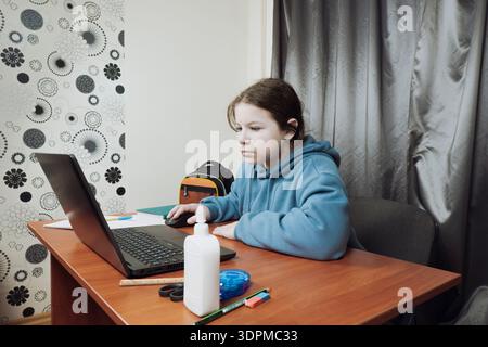 Écolière assise à un bureau en bois et utilisant un ordinateur portable pour l'apprentissage à distance ou les devoirs à la maison. Outils pédagogiques et sac à dos sur la table Banque D'Images