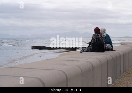 Deux amies anonymes assises sur un rebord passant du temps ensemble et partageant un moment à la plage de Strand, Cape Town, Afrique du Sud. Banque D'Images