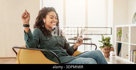 Femme utilisant un smartphone et portant des écouteurs tout en étant assis dans le salon Banque D'Images