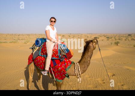 Jaisalmer, Inde - 29 mars 2022 : une personne portant des lunettes de soleil et des vêtements décontractés est assise sur un chameau avec une selle colorée dans le paysage sablonneux du désert du Thar sous un ciel dégagé. Banque D'Images