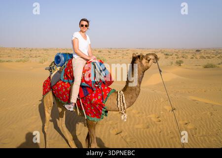 Jaisalmer, Inde - 29 mars 2022 : une personne portant des lunettes de soleil et des vêtements décontractés est assise sur un chameau avec une selle colorée et un récipient d'eau dans le paysage sablonneux du désert du Thar sous un ciel dégagé. Banque D'Images